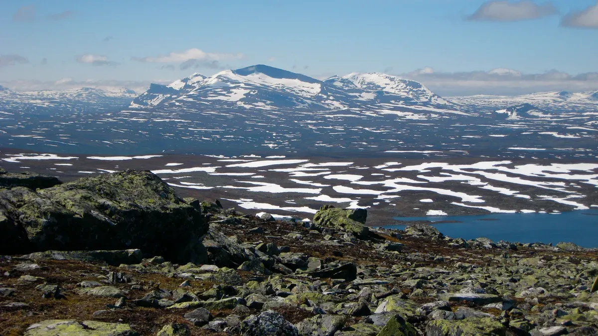 Vy från fjällterrängen ovanför Helags med snöfläckar, sjö och fjällkedjan i bakgrunden
