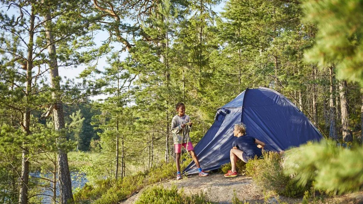 Två personer slår upp ett blått tält i skogen nära en sjö