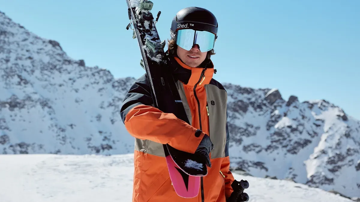 En vandrare i en freerideoverall med hjälm bär skidor över axeln framför en bergstopp