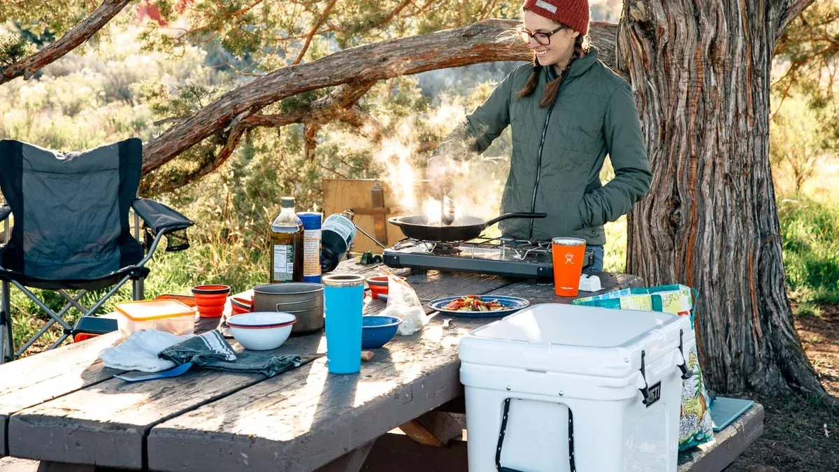 Campingkök på ett bord med stormkök, stekpanna, kylbox och köksredskap utomhus