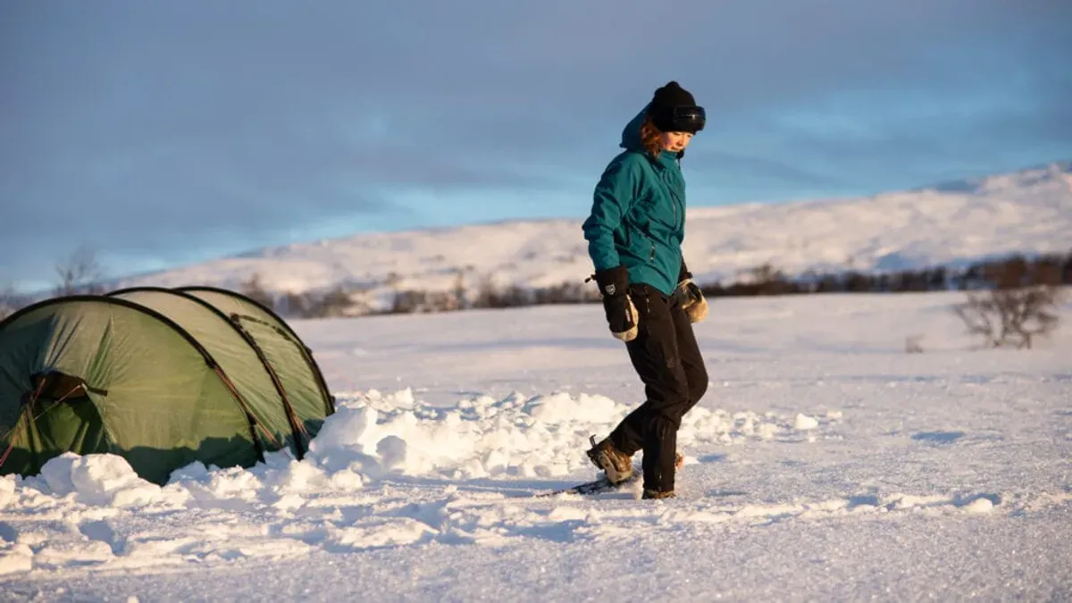 Vandrare i snöskor går förbi ett grönt tält i snölandskap i lågt vintersolljus