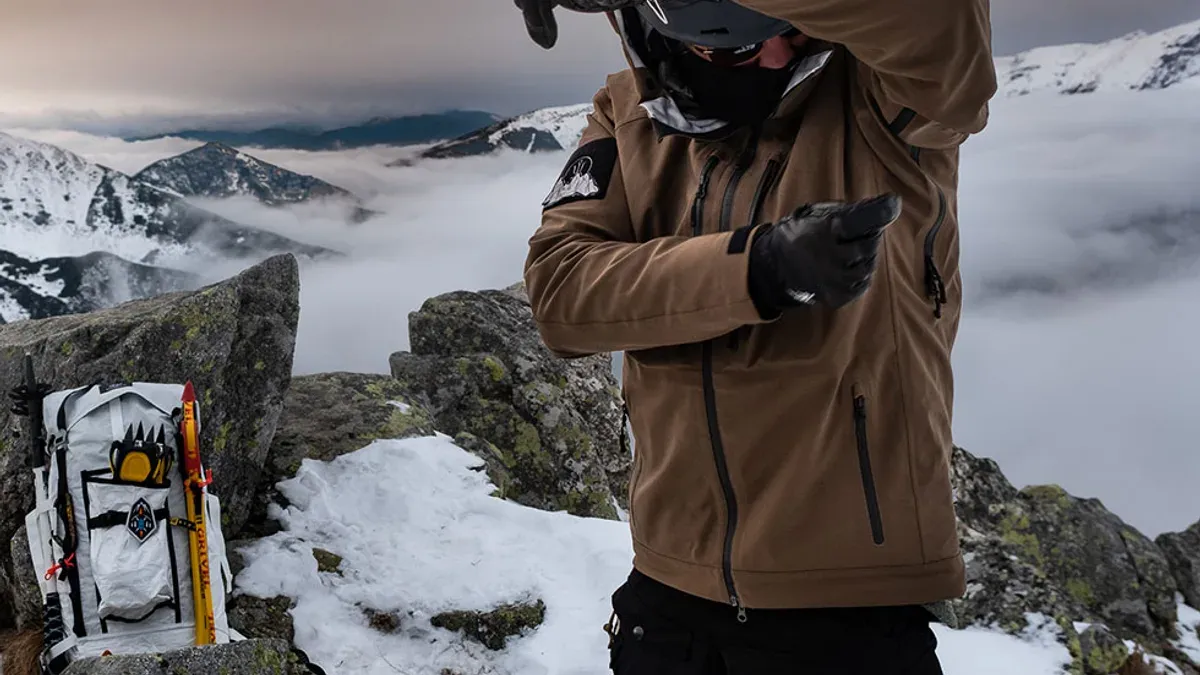 Man i brun skaljacka och svarta handskar på bergstopp
