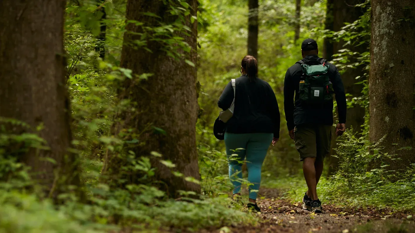 Två vandrare med ryggsäckar går genom en grön skog på en skogsstig