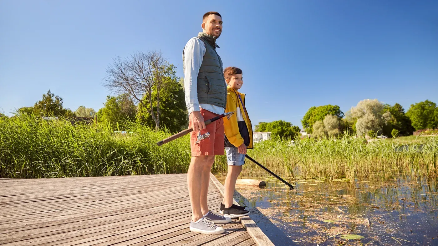 En pappa och en son står på en brygga med varsitt fiskespö en solig sommardag