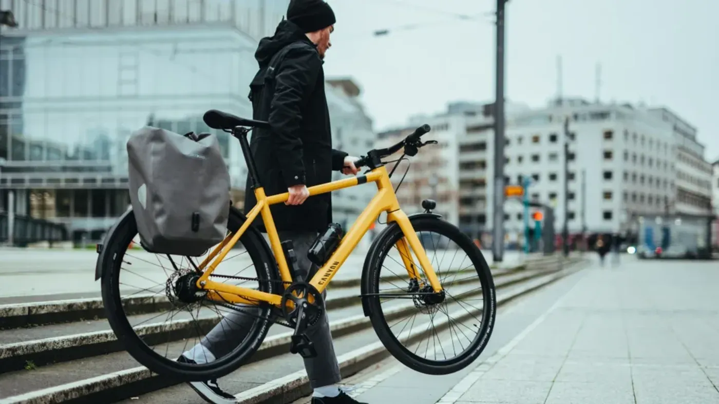 Cyklist i mörka kläder bär en gul hybridcykel nedför en trappa i en stadsmiljö