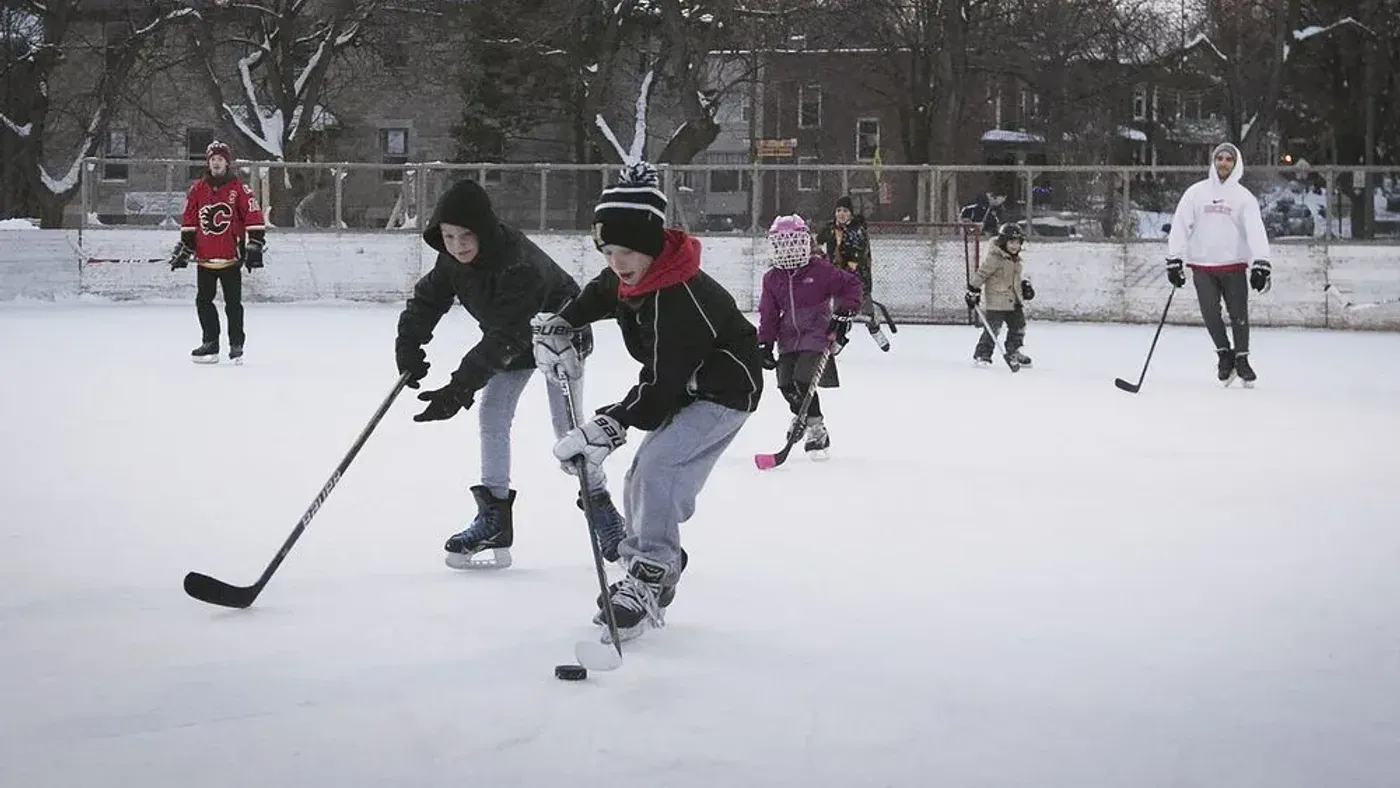Barn och ungdomar spelar ishockey på en utomhusrink med snö och trähus i bakgrunden