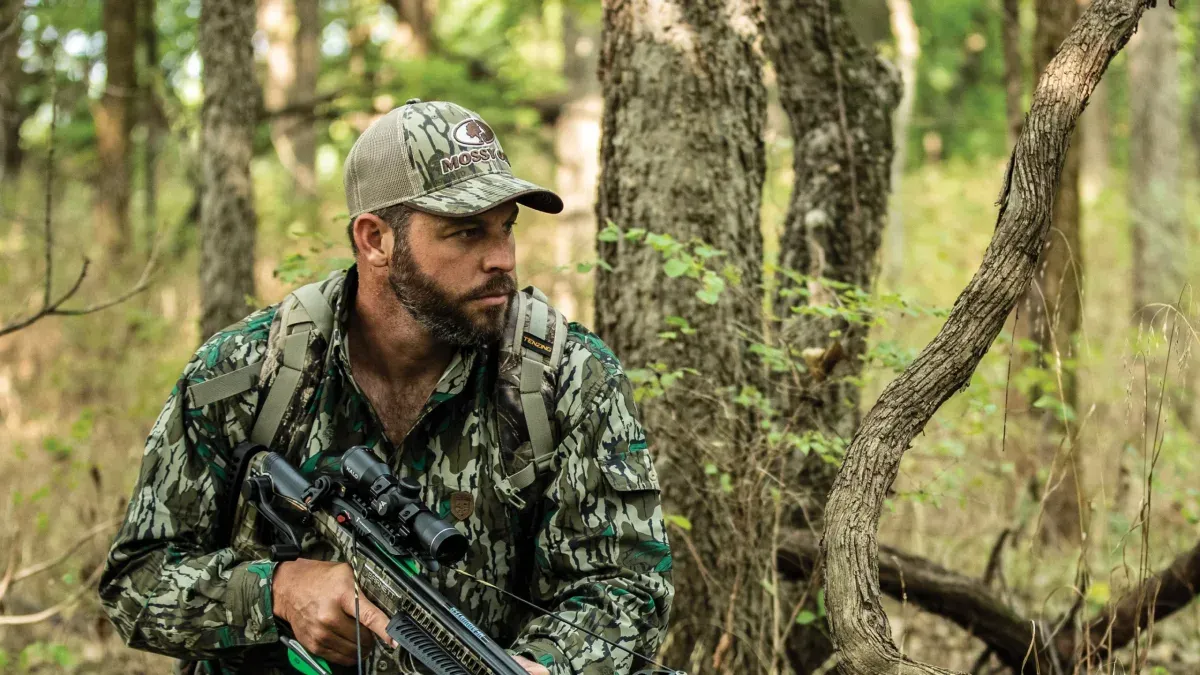 Jägare i kamouflagejacka med kikarsiktat vapen i skogen