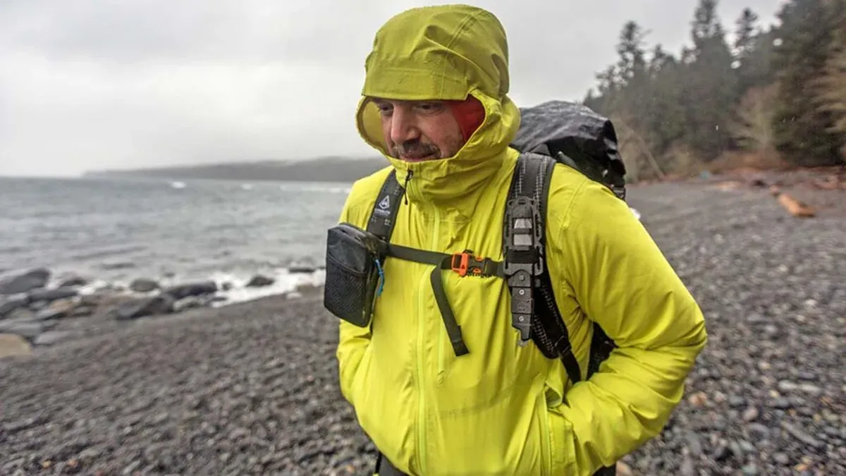 Man i gulgrön skaljacka på klippig strand med ryggsäck