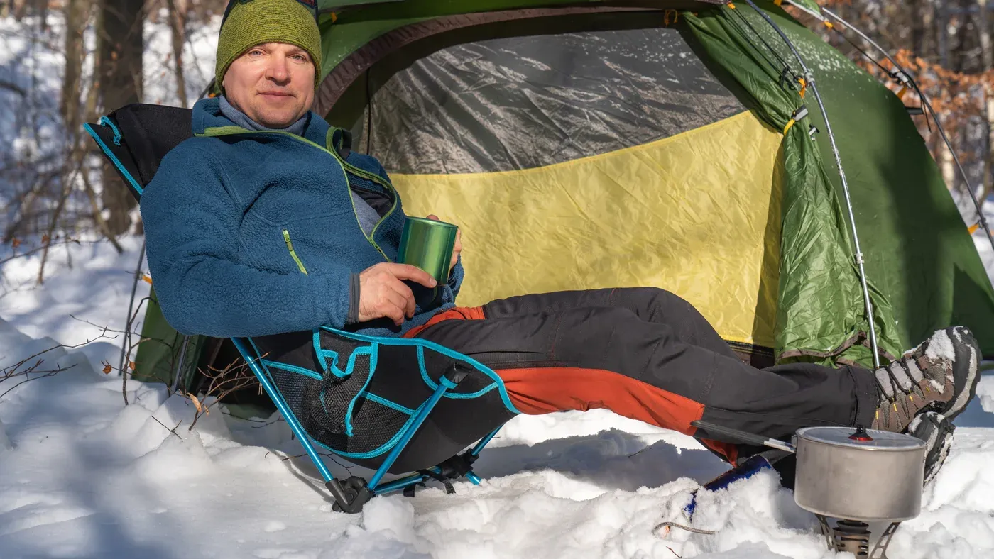 Man sitter i campingstol utanför tält med mugg i handen