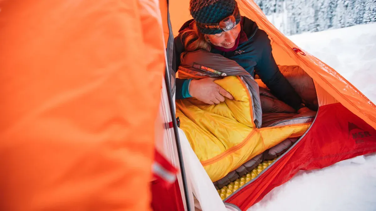Kvinna packar ner gul vintersovsäck i orange tält i snön
