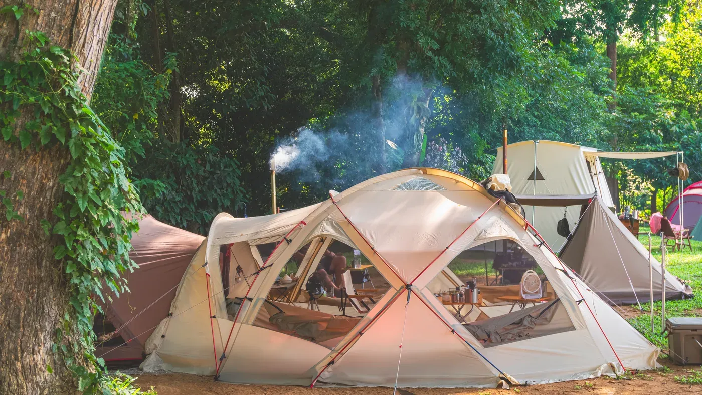 Stort beige campingtält med rökande skorsten bland träd