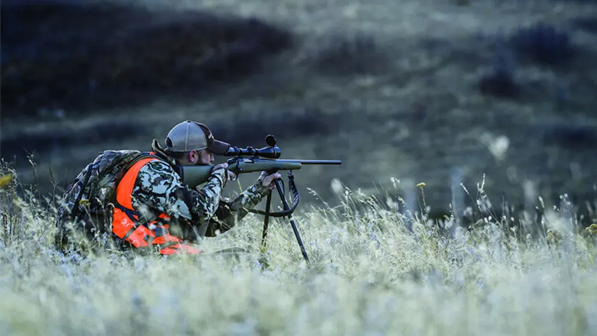 Jägare i orange varselväst siktar med stöd i högt gräs