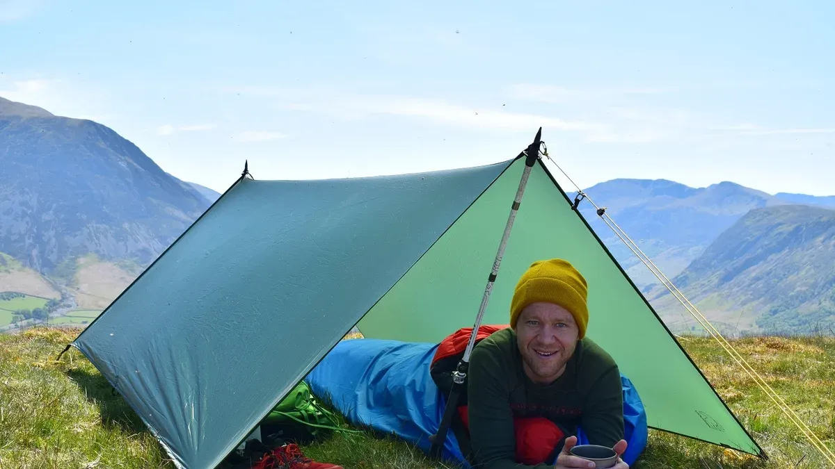 Man ligger under grön tarp i fjällterräng med kaffekopp i handen