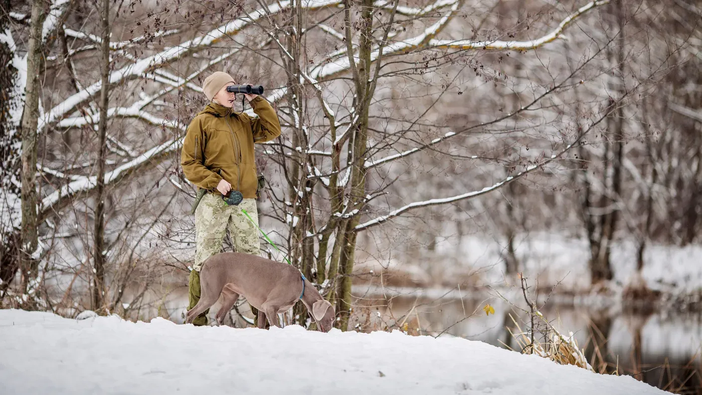 Jägare i termobyxor med hund i koppel i snötäckt skog