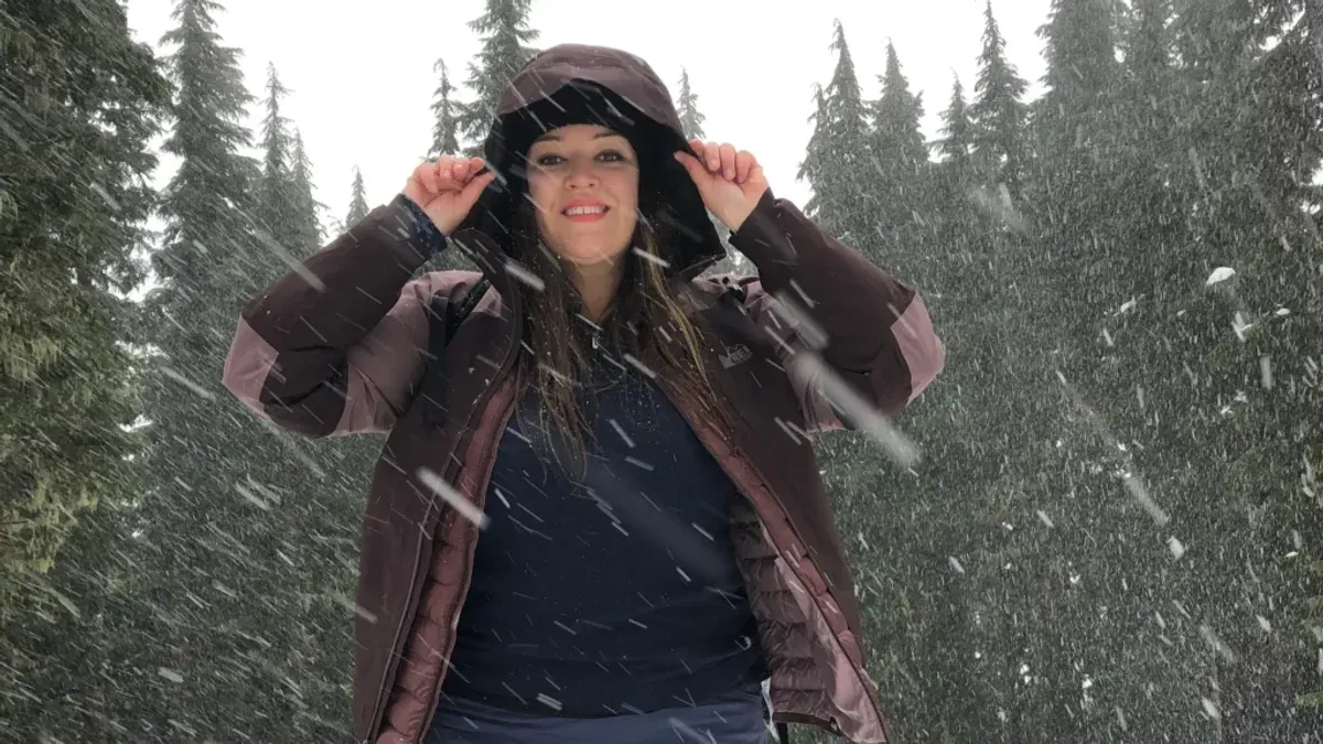 Kvinna drar upp luvan på brun vinterjacka i snöfall