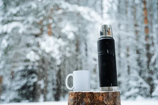 Svart termos och vit mugg på trädstubbe i snöfall