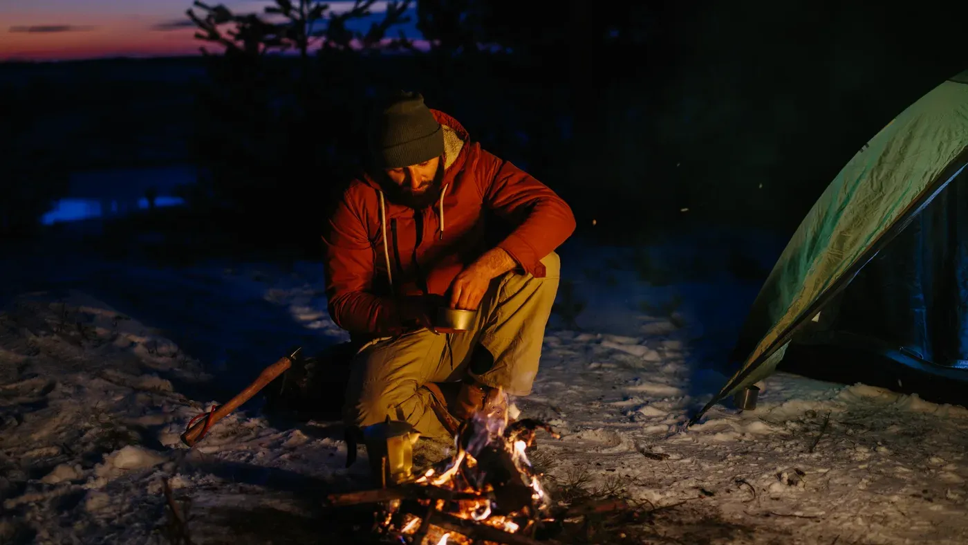 Man vid lägereld i mörker med tält och snö i bakgrunden