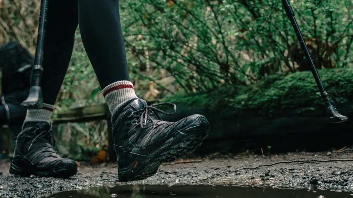 Fötter i vandringsskor och stavar vid vattenpöl i skog