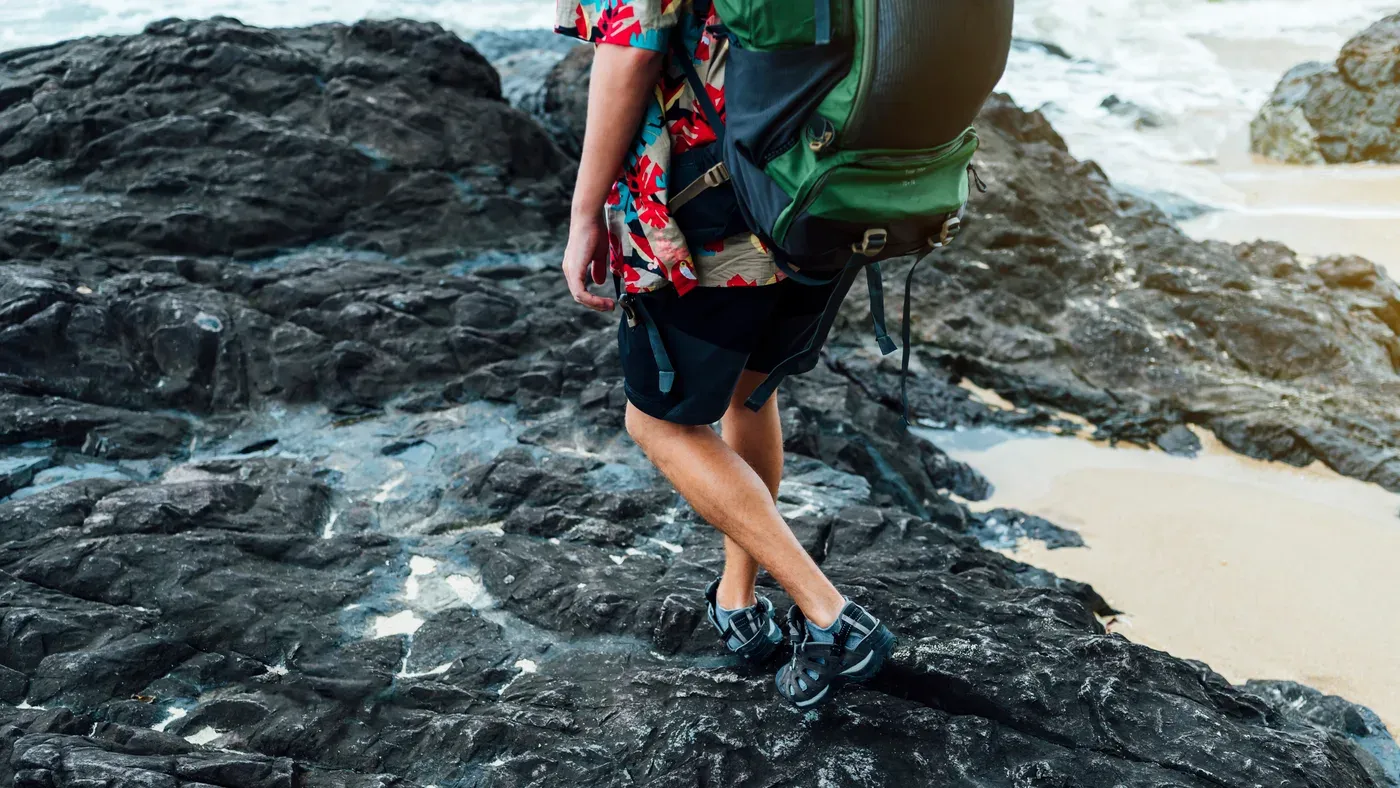 Person i shorts och hawaiiskjorta med ryggsäck på klippor vid stranden